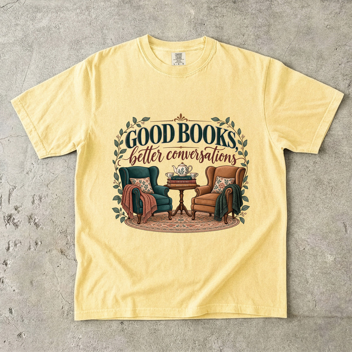 Butter T-shirt featuring “Good Books, Better Conversations” with two armchairs around a tea table, stacked books, floral cushions and leafy framing.