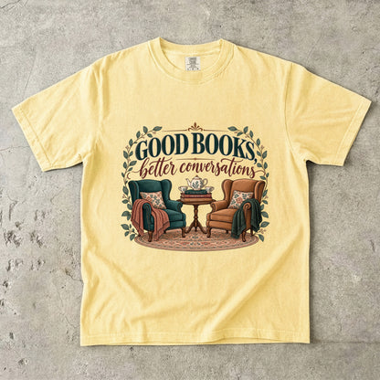 Butter T-shirt featuring “Good Books, Better Conversations” with two armchairs around a tea table, stacked books, floral cushions and leafy framing.