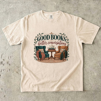Ivory T-shirt featuring “Good Books, Better Conversations” with two armchairs around a tea table, stacked books, floral cushions and leafy framing.