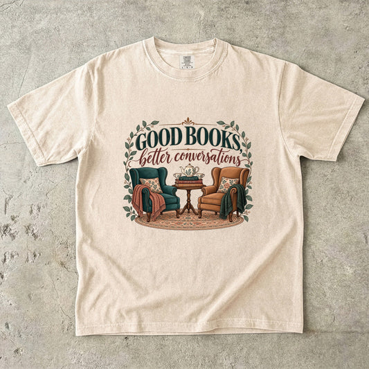 Ivory T-shirt featuring “Good Books, Better Conversations” with two armchairs around a tea table, stacked books, floral cushions and leafy framing.