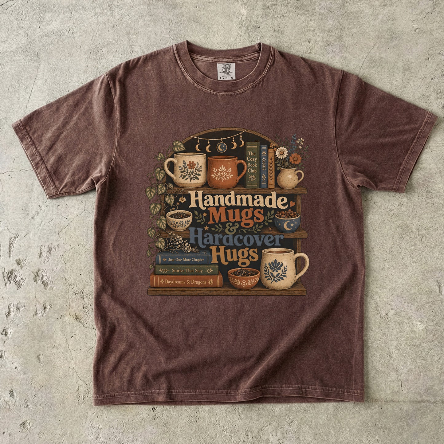 Front view of an Espresso Comfort Colors book lover shirt featuring handmade mugs, hardcover books, botanical shelves and the phrase "Handmade Mugs & Hardcover Hugs