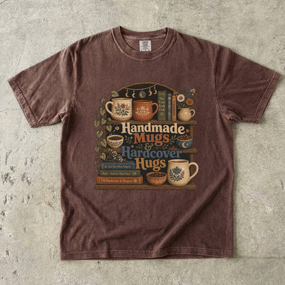 Front view of an Espresso Comfort Colors book lover shirt featuring handmade mugs, hardcover books, botanical shelves and the phrase "Handmade Mugs & Hardcover Hugs