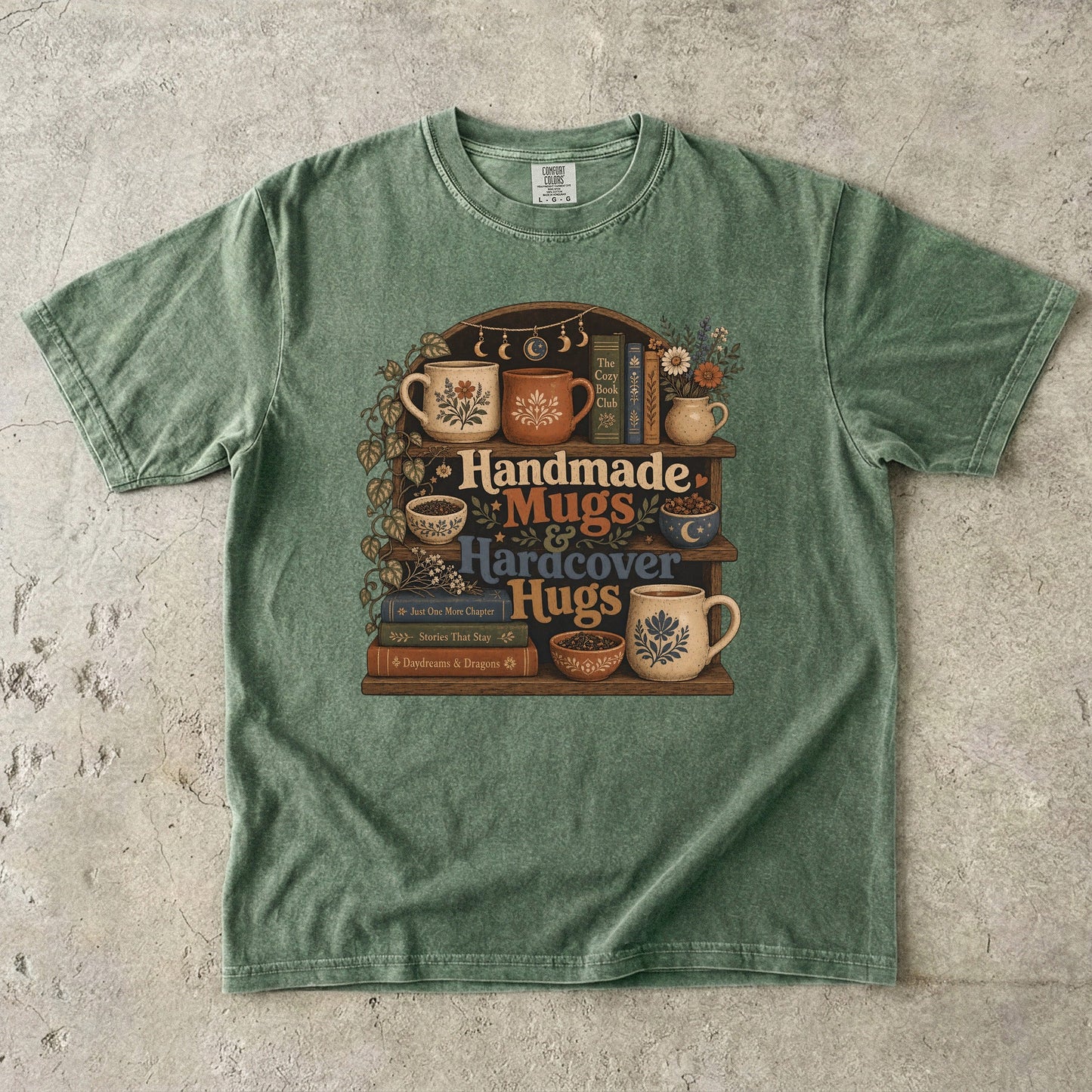Front view of a Moss Comfort Colors book lover shirt featuring handcrafted mugs, hardcover books and botanical bookshelf illustration.