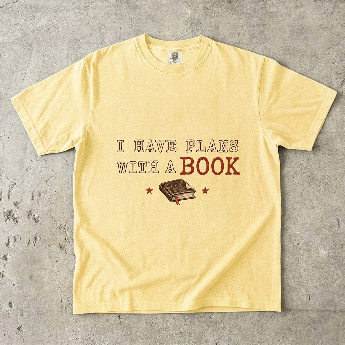 Butter T-shirt featuring “I Have Plans With a Book” with distressed cream and rust lettering, a closed book with a ribbon bookmark and two stars.