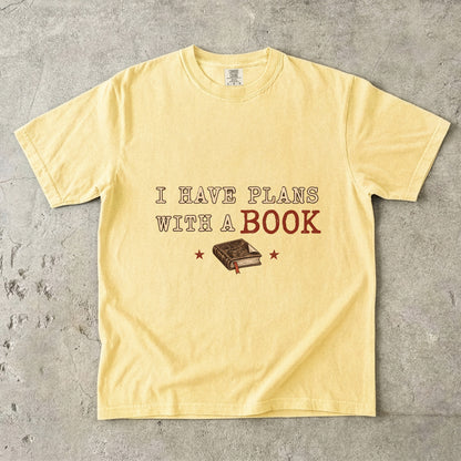 Butter T-shirt featuring “I Have Plans With a Book” with distressed cream and rust lettering, a closed book with a ribbon bookmark and two stars.