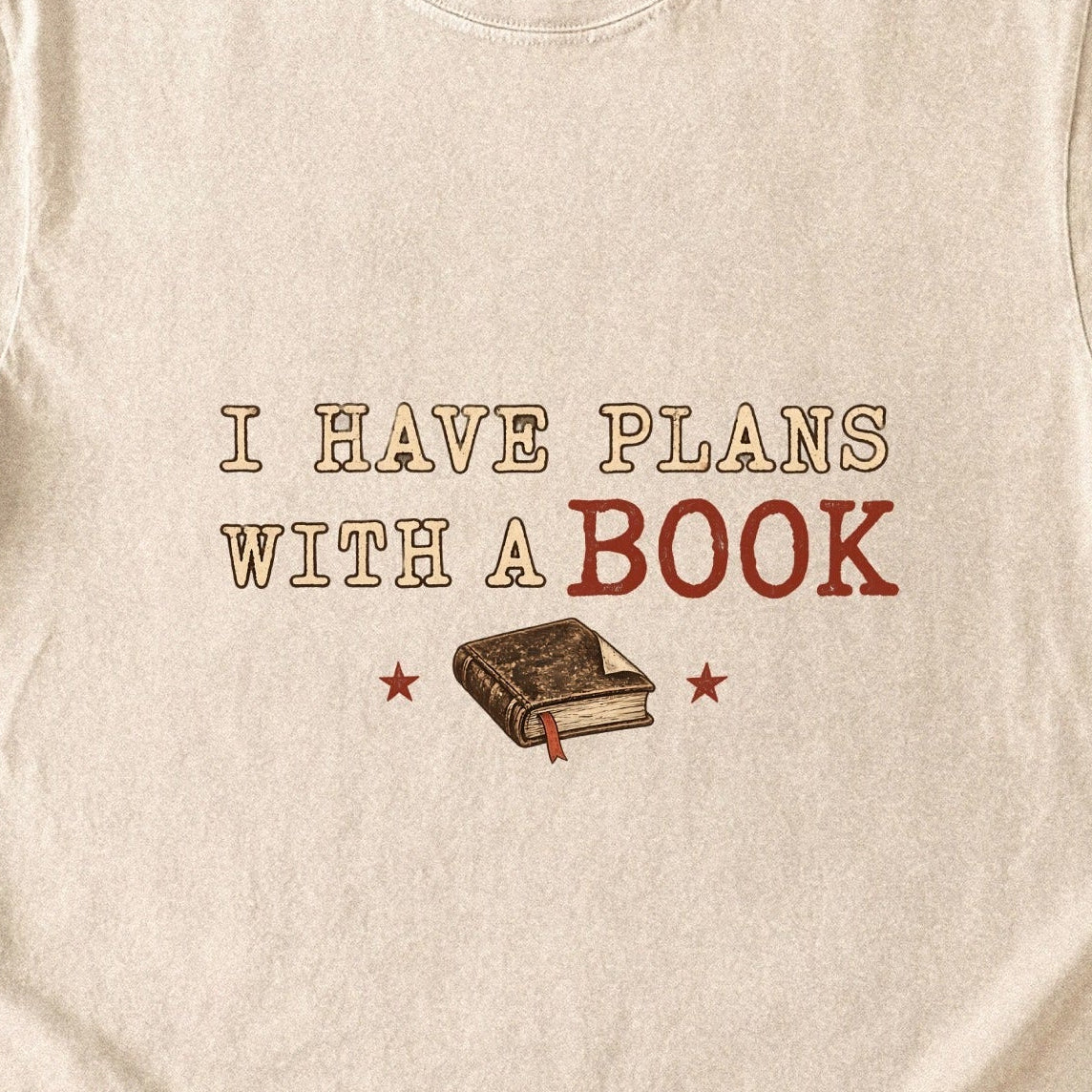 Close-up of the “I Have Plans With a Book” T-shirt design, showing distressed cream and rust lettering, a closed book with a ribbon bookmark and two stars.