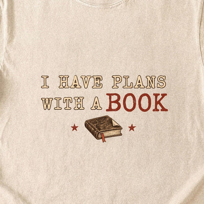 Close-up of the “I Have Plans With a Book” T-shirt design, showing distressed cream and rust lettering, a closed book with a ribbon bookmark and two stars.