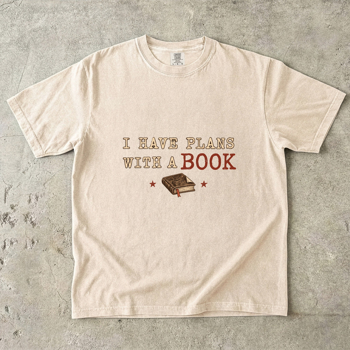 Ivory T-shirt featuring “I Have Plans With a Book” with distressed cream and rust lettering, a closed book with a ribbon bookmark and two stars.