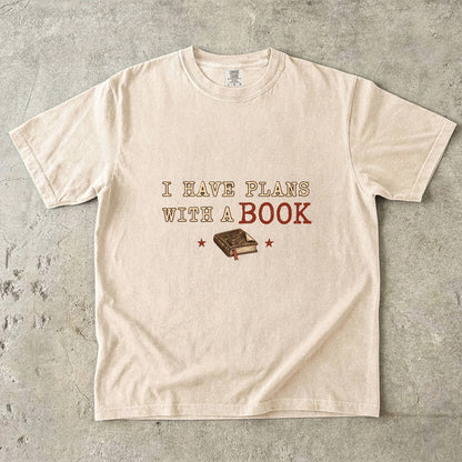 Ivory T-shirt featuring “I Have Plans With a Book” with distressed cream and rust lettering, a closed book with a ribbon bookmark and two stars.