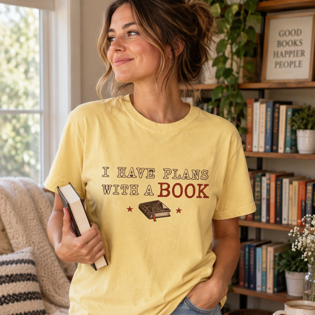 A woman wearing a pale yellow butter graphic t-shirt that reads 'I HAVE PLANS WITH A BOOK' with a small book illustration, paired with denim shorts, standing in a cozy, book-filled room.