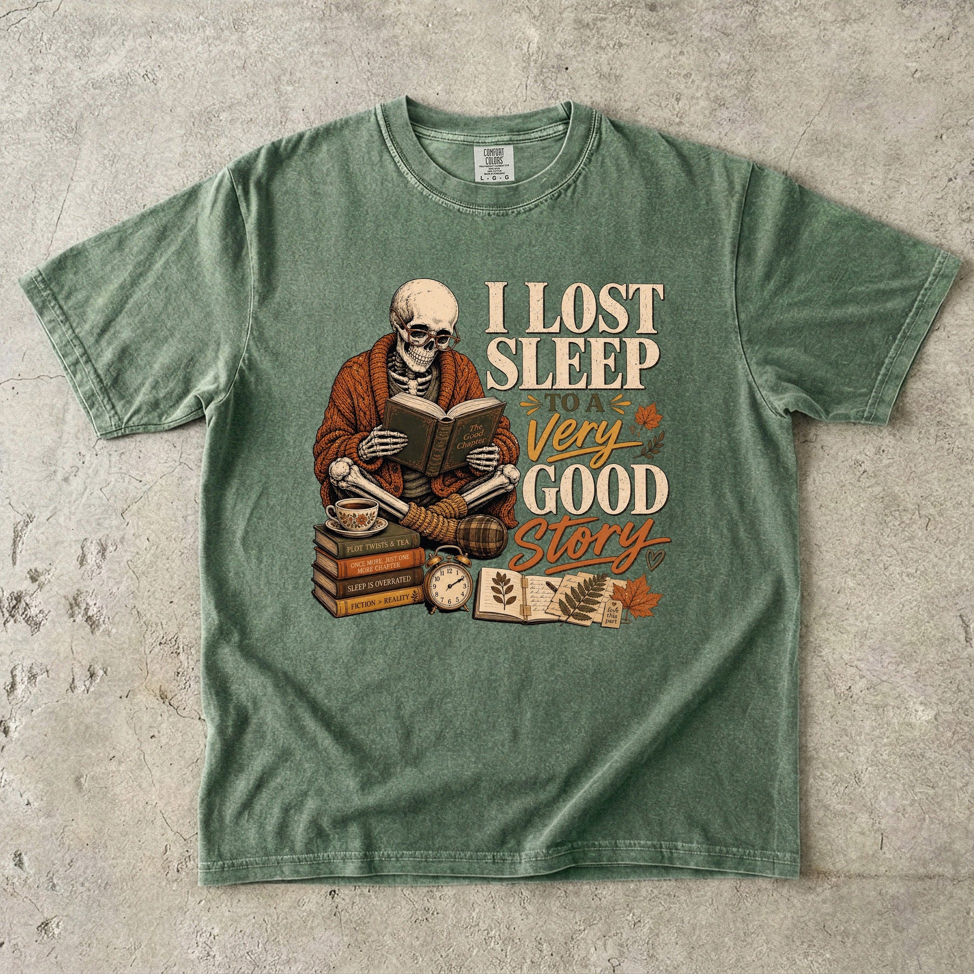 Moss Comfort Colors book lover T-shirt featuring a skeleton reading beside stacked books, tea and a clock.