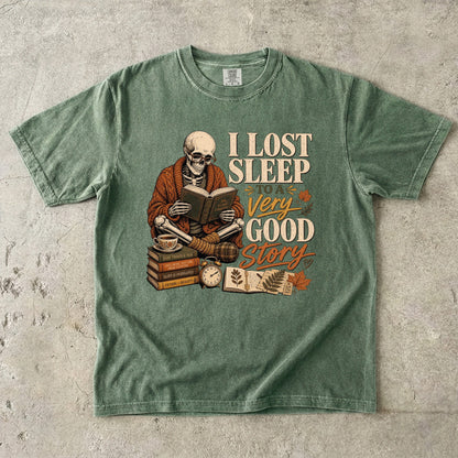 Moss Comfort Colors book lover T-shirt featuring a skeleton reading beside stacked books, tea and a clock.