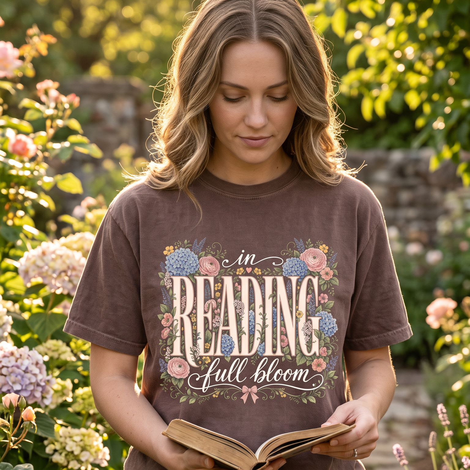 Reader holding an open book while wearing the In Reading, Full Bloom floral book lover T-shirt in a sunlit flower garden.