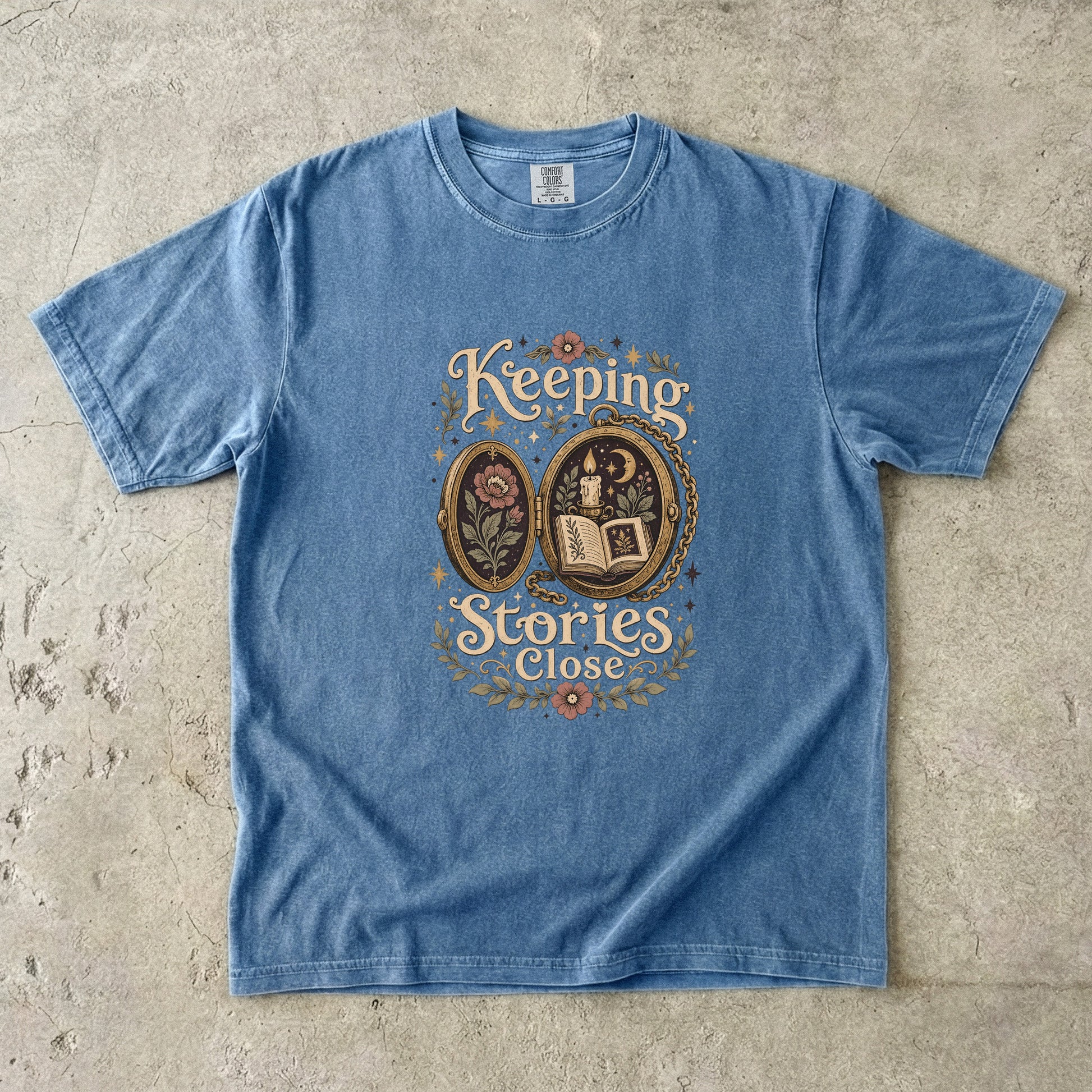 Front view of a Blue Jean Comfort Colors Book Lover T-shirt with the Keeping Stories Close literary illustration.