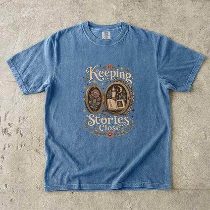 Front view of a Blue Jean Comfort Colors Book Lover T-shirt with the Keeping Stories Close literary illustration.