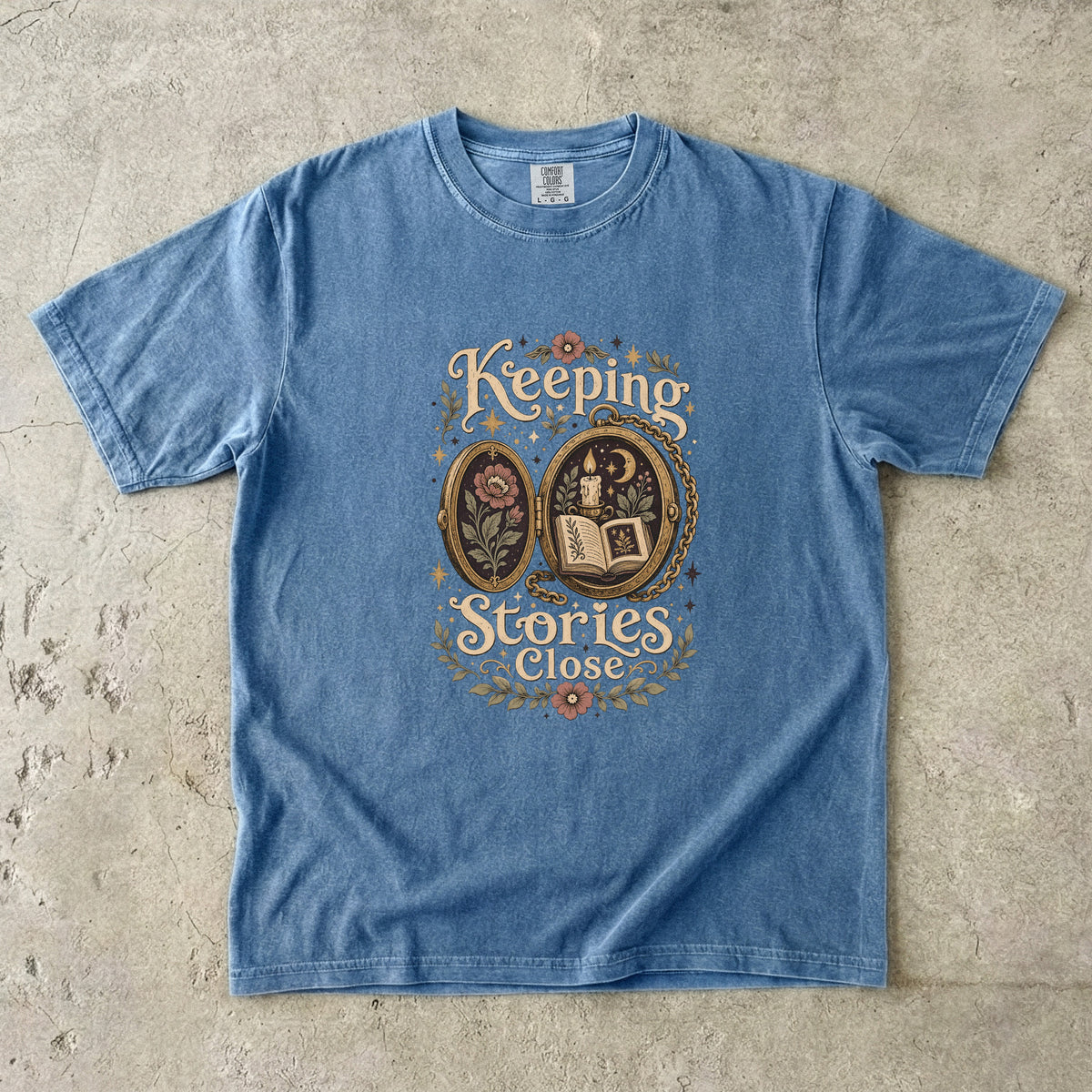 Keeping Stories Close book lover T-shirt in Blue Jean featuring an ornate locket with an open book, candle, moon and flowers