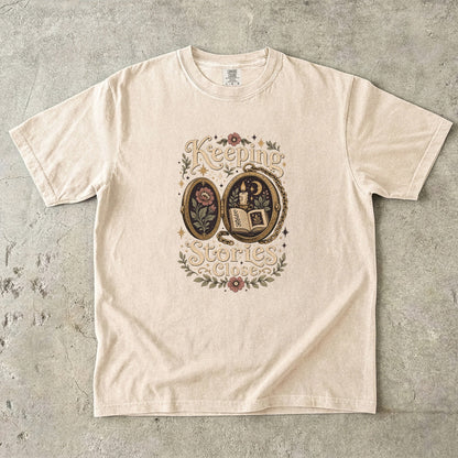 Front view of an Ivory Comfort Colors Book Lover T-shirt featuring the Keeping Stories Close vintage literary locket illustration.