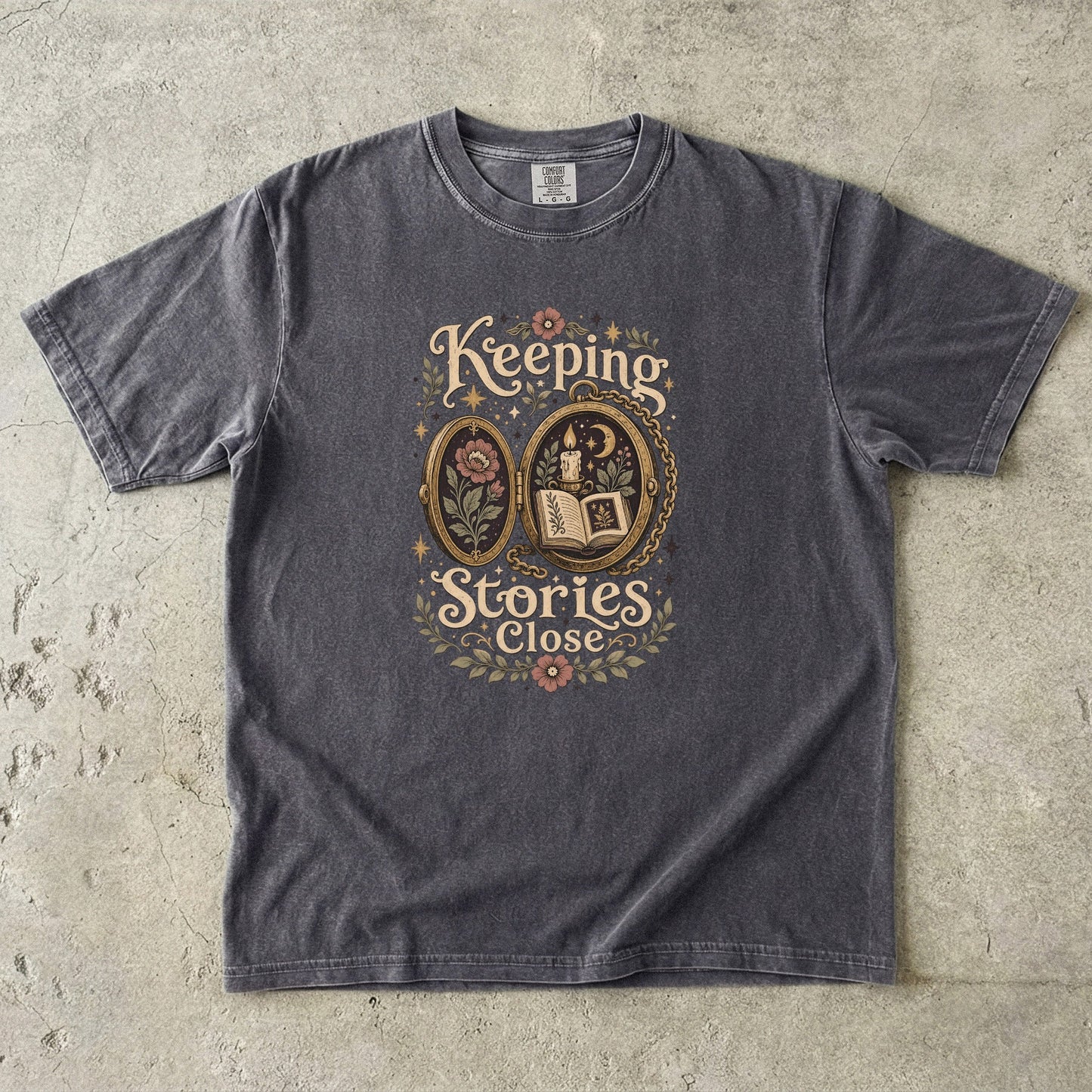 Front view of a Pepper Comfort Colors Book Lover T-shirt featuring the Keeping Stories Close vintage storybook locket illustration with an open book, candlelight and botanical details.