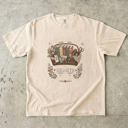 Mending Hearts With Stories book club T-shirt in Ivory featuring classic books, sewing notions, flowers and a woven basket
