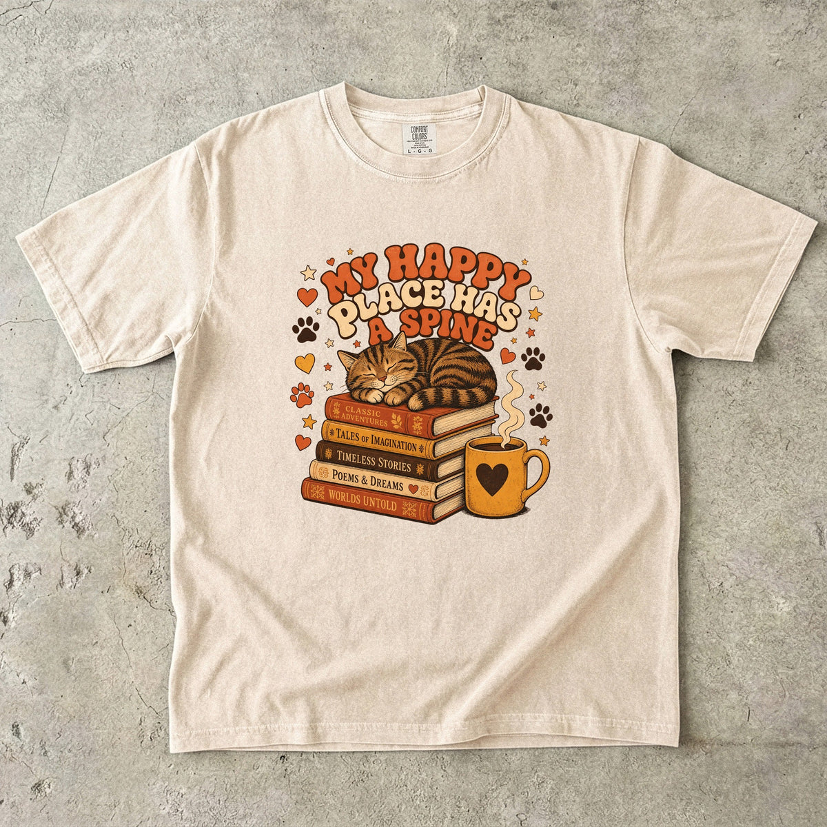 Ivory cat book lover T-shirt featuring a sleeping tabby on stacked books beside a steaming mug with hearts, stars and paw prints.