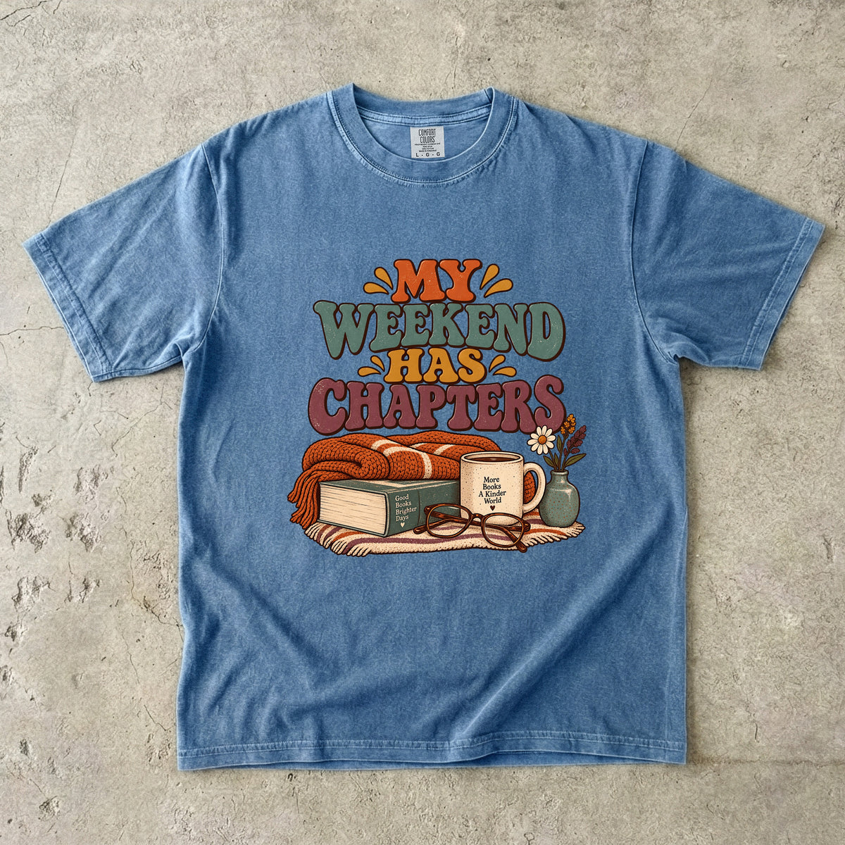 Blue Jean T-shirt featuring “My Weekend Has Chapters” with multicolored vintage lettering, a knitted blanket, book, mug, glasses and a small flower vase.