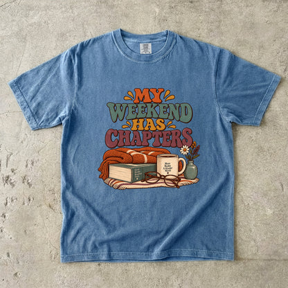 Blue Jean T-shirt featuring “My Weekend Has Chapters” with multicolored vintage lettering, a knitted blanket, book, mug, glasses and a small flower vase.