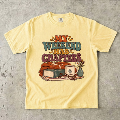 Butter T-shirt featuring “My Weekend Has Chapters” with multicolored vintage lettering, a knitted blanket, book, mug, glasses and a small flower vase.