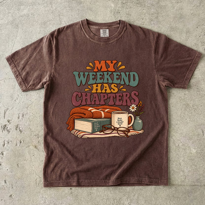 Espresso T-shirt featuring “My Weekend Has Chapters” with multicolored vintage lettering, a knitted blanket, book, mug, glasses and a small flower vase.