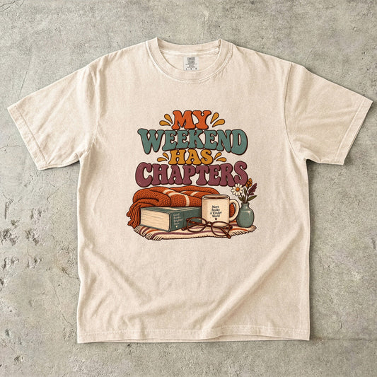 Ivory T-shirt featuring “My Weekend Has Chapters” with multicolored vintage lettering, a knitted blanket, book, mug, glasses and a small flower vase.