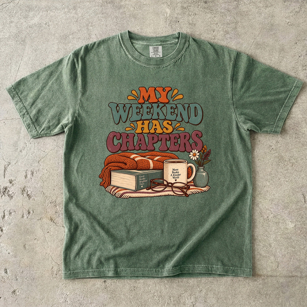 Moss T-shirt featuring “My Weekend Has Chapters” with multicolored vintage lettering, a knitted blanket, book, mug, glasses and a small flower vase.