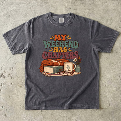 Pepper T-shirt featuring “My Weekend Has Chapters” with multicolored vintage lettering, a knitted blanket, book, mug, glasses and a small flower vase.