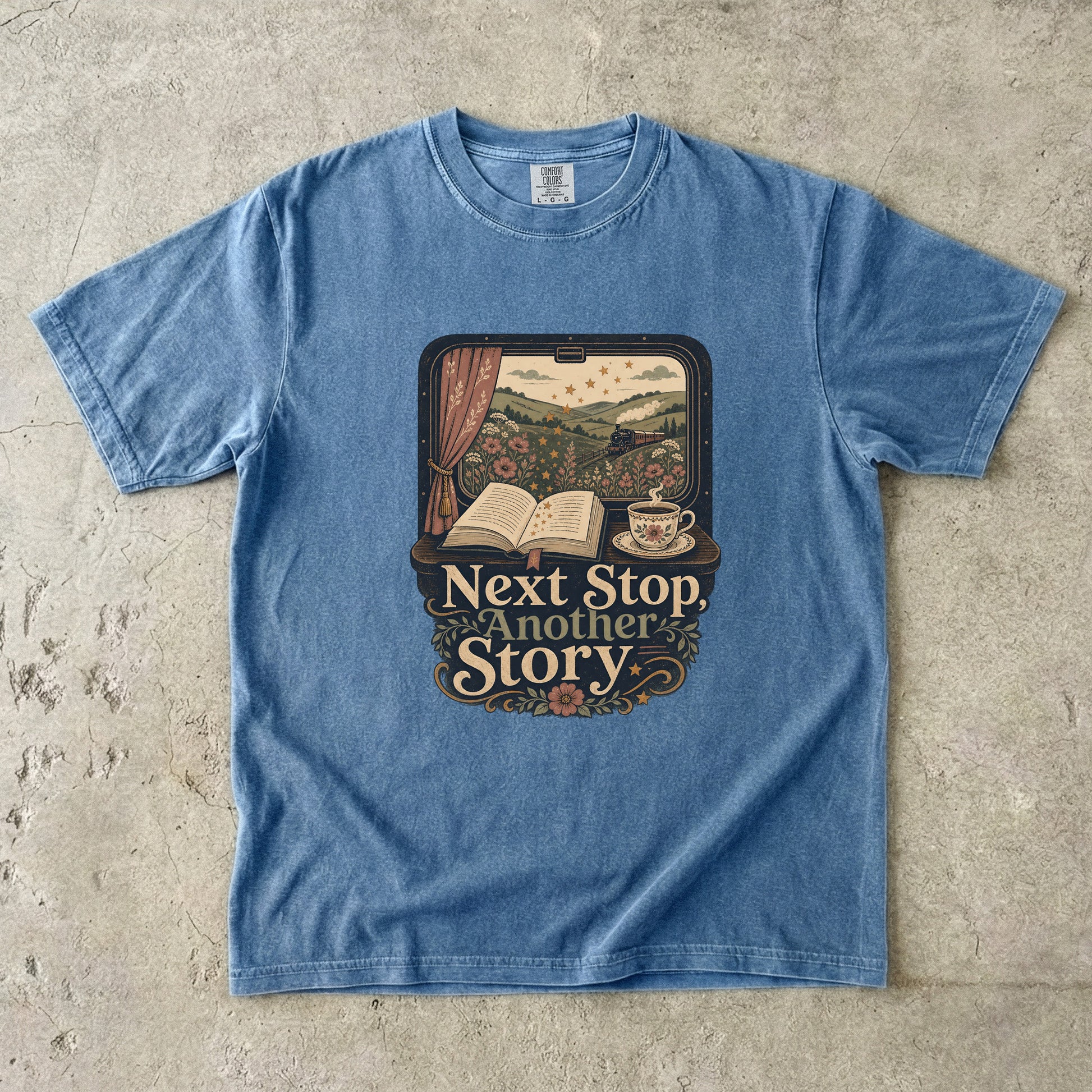 Front view of a Blue Jean Comfort Colors Book Lover Shirt featuring a vintage train window, open book, teacup and the phrase "Next Stop, Another Story."