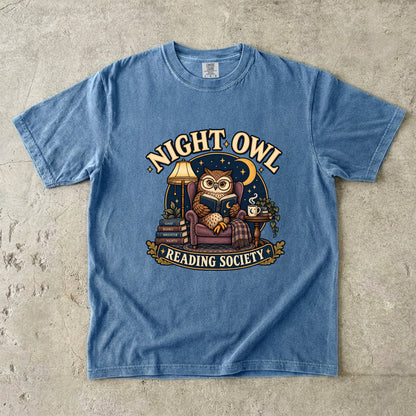 Blue Jean T-shirt featuring “Night Owl Reading Society” with a spectacled owl reading in an armchair, a lamp, books, mug, crescent moon and stars.