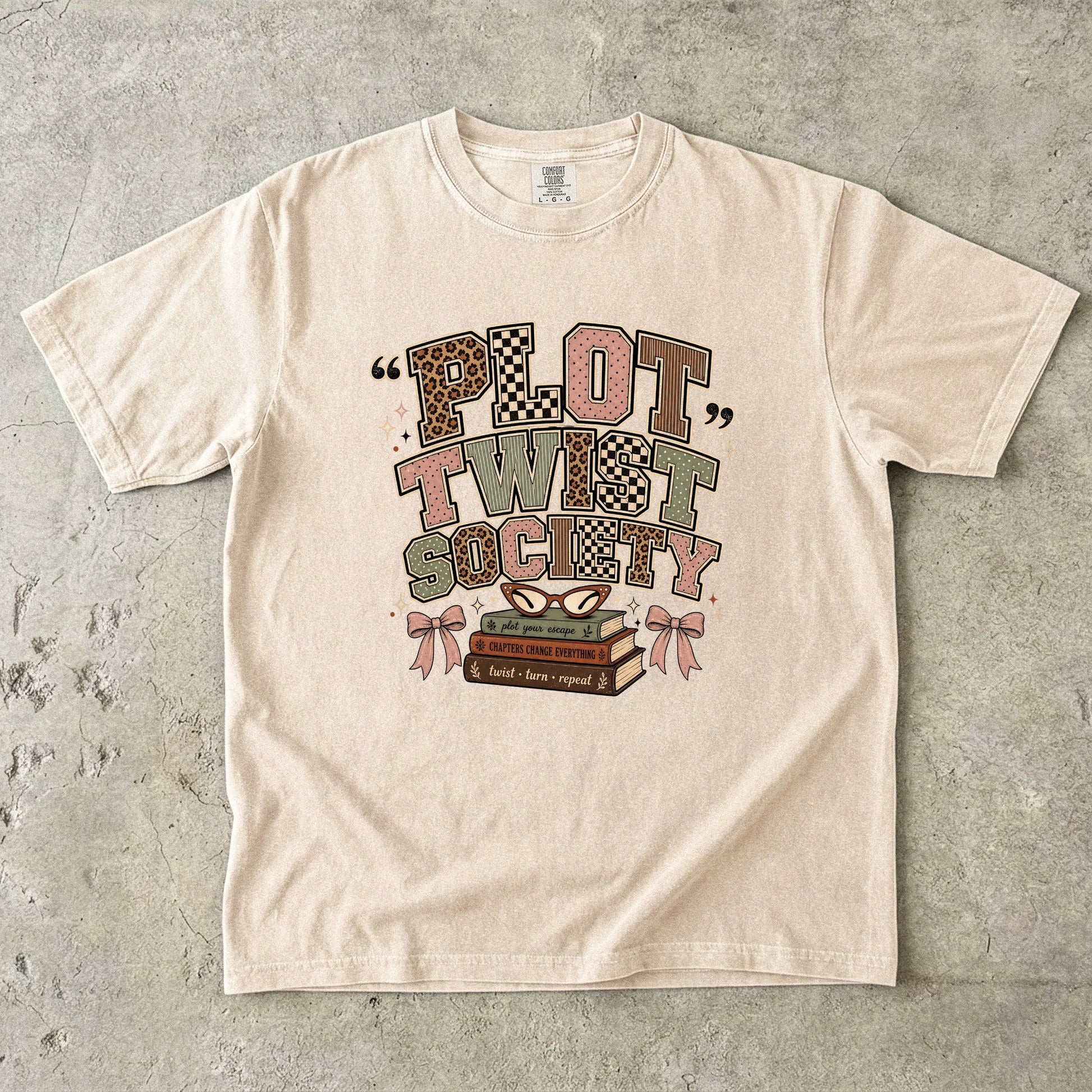 Ivory Comfort Colors Plot Twist Society T-shirt featuring patterned typography, stacked books, bows and reading glasses.
