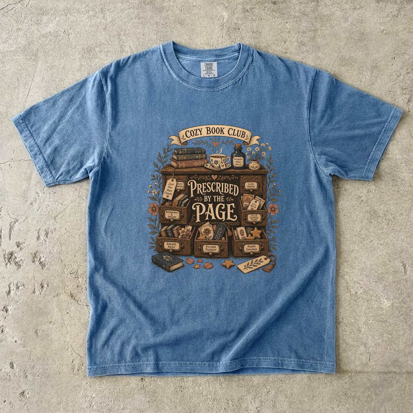 Prescribed by the Page cozy book club T-shirt in Blue Jean featuring a vintage literary cabinet filled with books, tea and reading-themed drawers