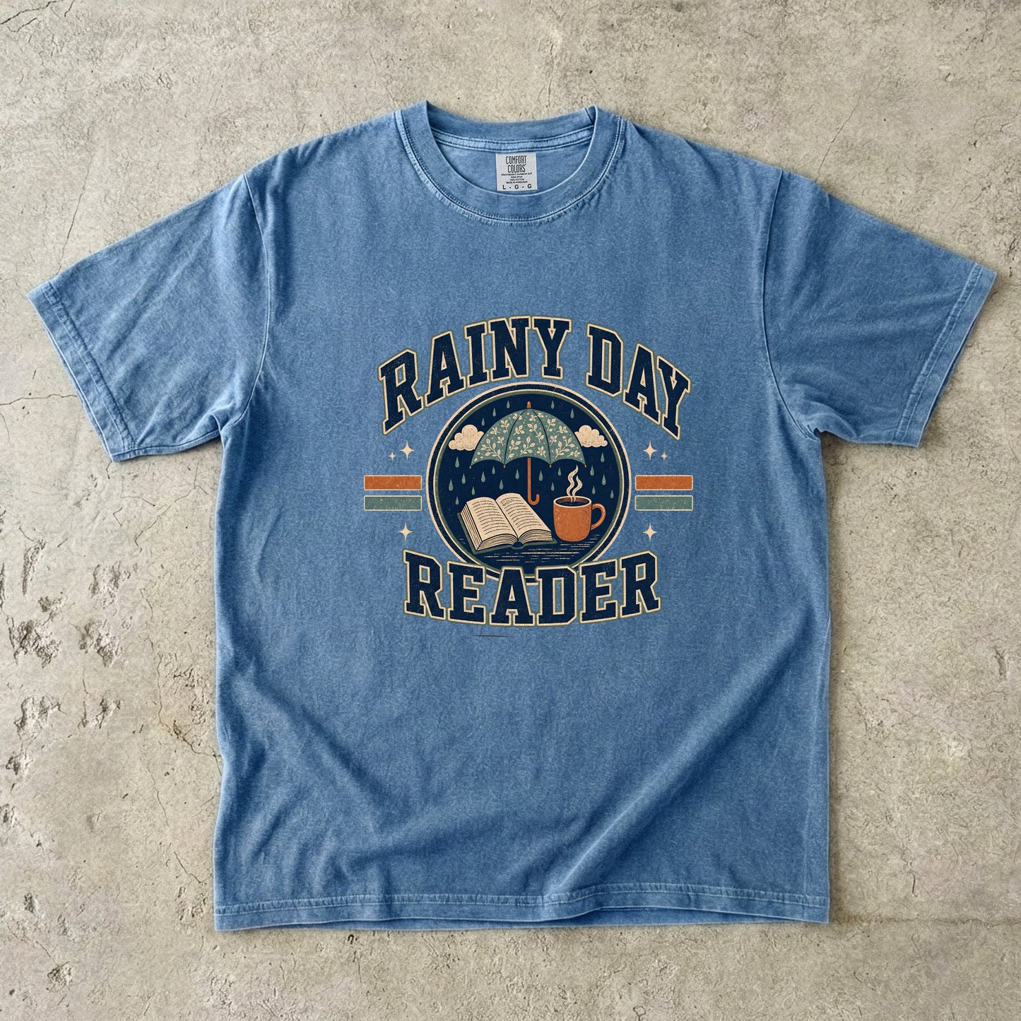 Blue Jean Comfort Colors book lover T-shirt featuring a Rainy Day Reader design with an umbrella, open book and warm drink.