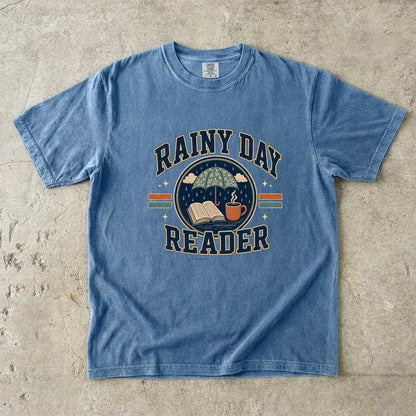 Blue Jean Comfort Colors book lover T-shirt featuring a Rainy Day Reader design with an umbrella, open book and warm drink.