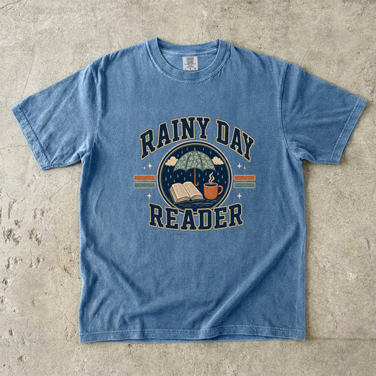Blue Jean Comfort Colors book lover T-shirt featuring a Rainy Day Reader design with an umbrella, open book and warm drink.