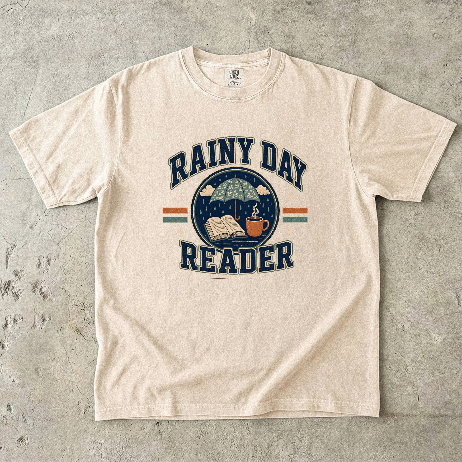Ivory Comfort Colors Rainy Day Reader T-shirt featuring a vintage umbrella, open book and warm drink illustration.