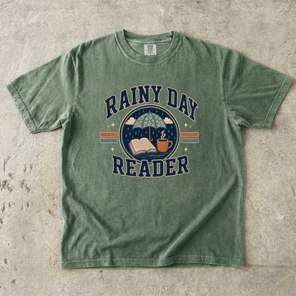 Ivory Comfort Colors Rainy Day Reader T-shirt featuring a vintage umbrella, open book and warm drink illustration.
