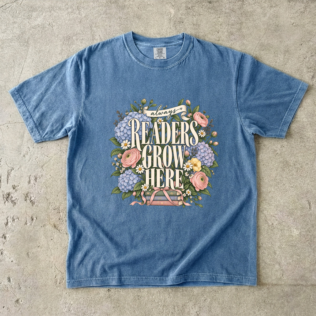 Blue Jean T-shirt featuring “always READERS GROW HERE” with large cream lettering surrounded by pastel blue flowers, pink blooms, daisies, leaves and ribbon-tied books.