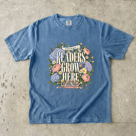 Blue Jean T-shirt featuring “always READERS GROW HERE” with large cream lettering surrounded by pastel blue flowers, pink blooms, daisies, leaves and ribbon-tied books.