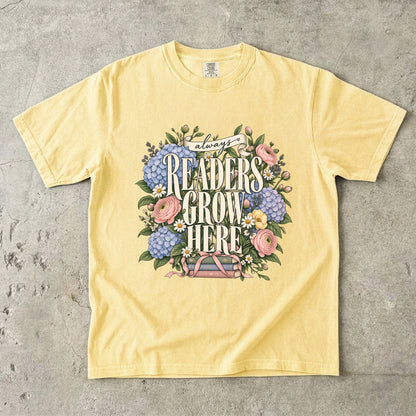 Butter T-shirt featuring “always READERS GROW HERE” with large cream lettering surrounded by pastel blue flowers, pink blooms, daisies, leaves and ribbon-tied books.