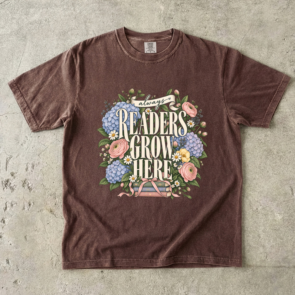 Espresso T-shirt featuring “always READERS GROW HERE” with large cream lettering surrounded by pastel blue flowers, pink blooms, daisies, leaves and ribbon-tied books.
