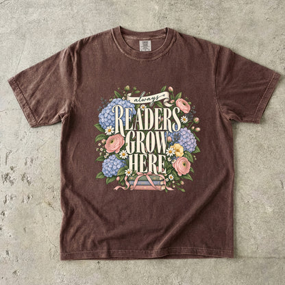Espresso T-shirt featuring “always READERS GROW HERE” with large cream lettering surrounded by pastel blue flowers, pink blooms, daisies, leaves and ribbon-tied books.