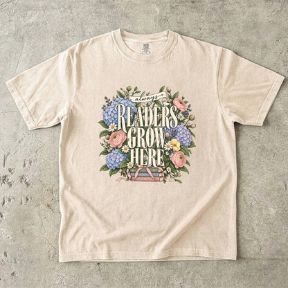 Ivory T-shirt featuring “always READERS GROW HERE” with large cream lettering surrounded by pastel blue flowers, pink blooms, daisies, leaves and ribbon-tied books.