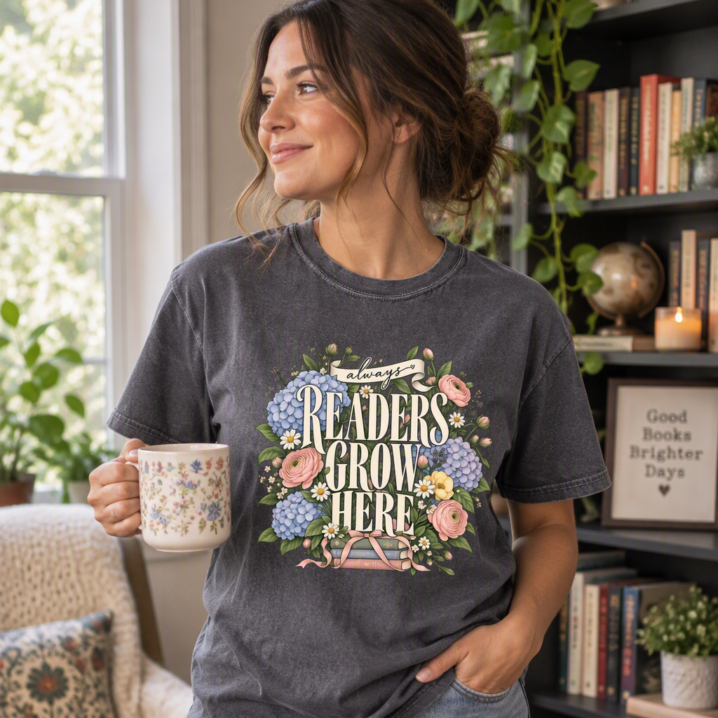 A woman wearing a pepper graphic t-shirt with a floral design that reads 'READERS GROW HERE' and 'Good Books Brighter Days', paired with light blue ripped jeans, holding a patterned mug in a cozy, book-filled room.