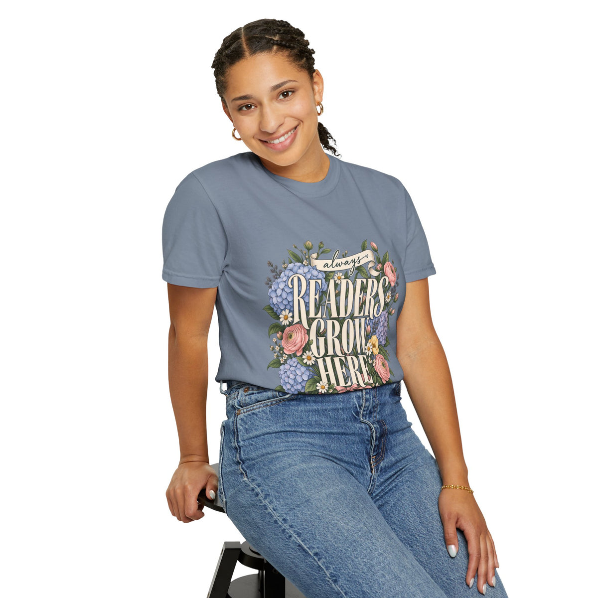 A woman wearing a slate blue jean graphic t-shirt with a floral design that reads 'always READERS GROW HERE' and blue jeans, posing against a solid white background.