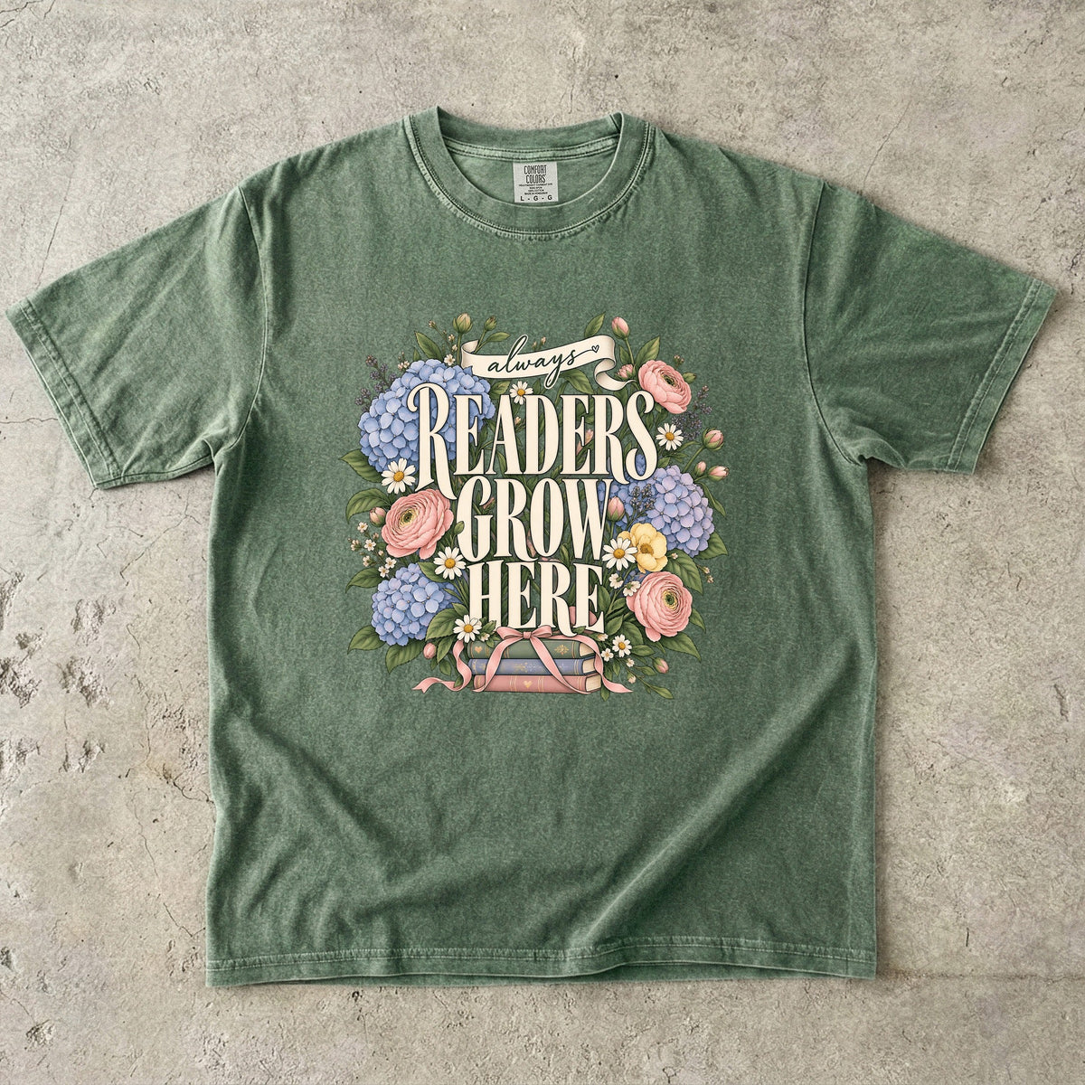 Moss T-shirt featuring “always READERS GROW HERE” with large cream lettering surrounded by pastel blue flowers, pink blooms, daisies, leaves and ribbon-tied books.