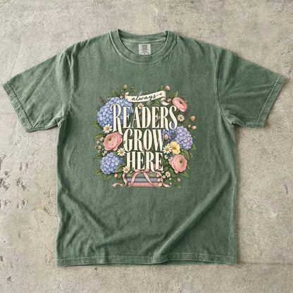 Moss T-shirt featuring “always READERS GROW HERE” with large cream lettering surrounded by pastel blue flowers, pink blooms, daisies, leaves and ribbon-tied books.